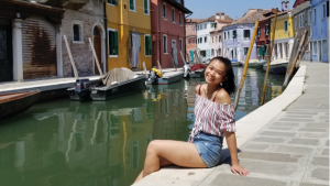 Thao Atkinson sitting along a canal in Venice in front of colourful buildings and boats.